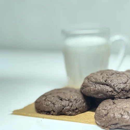 En masse brune chokolade småkager samlet, med et glas mælk bag dem, sat på et stykke bagepapir.